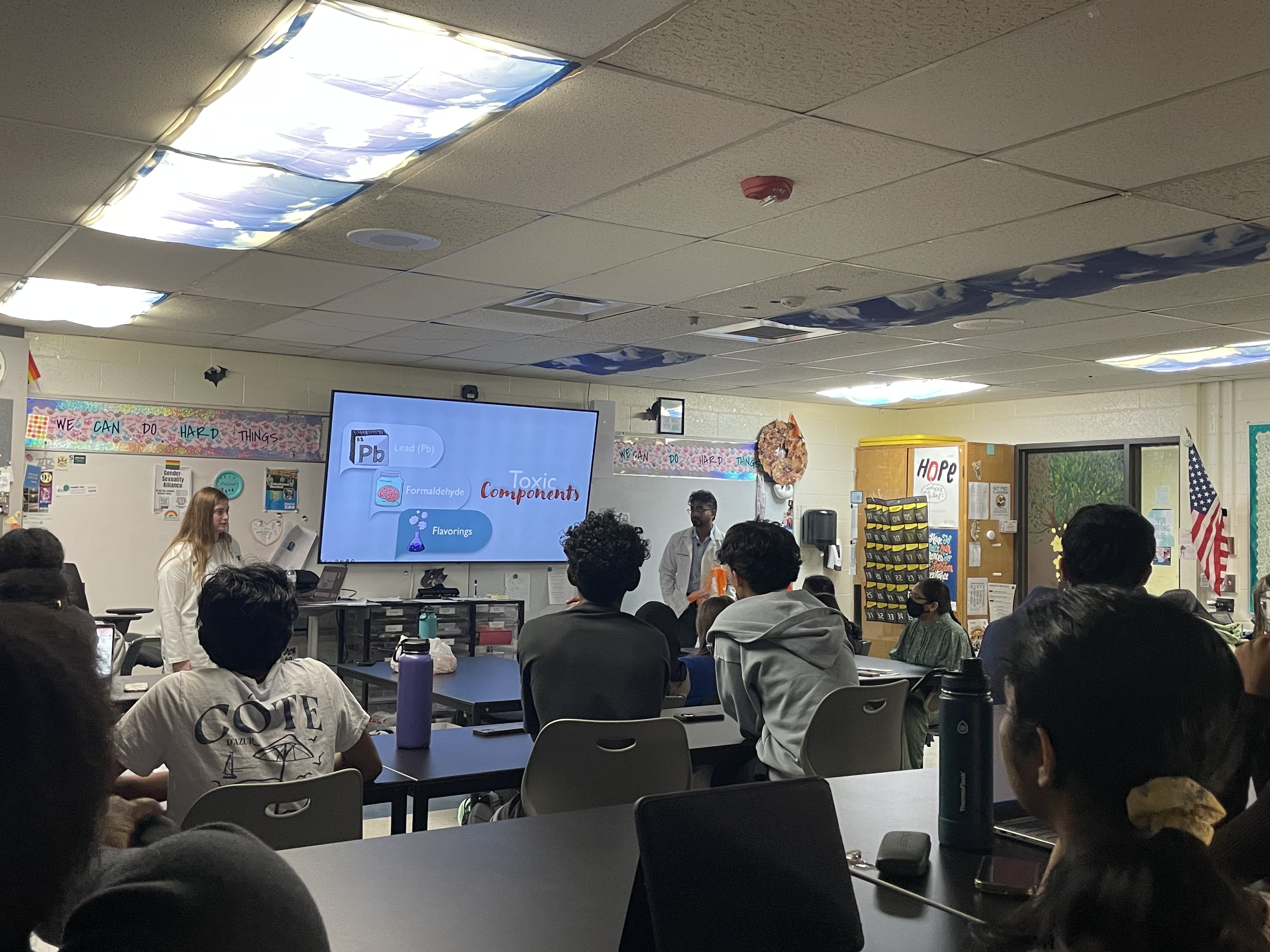 Students in a classroom facing forward looking at a large screen in the middle of the room showing a presentation titled toxic components..