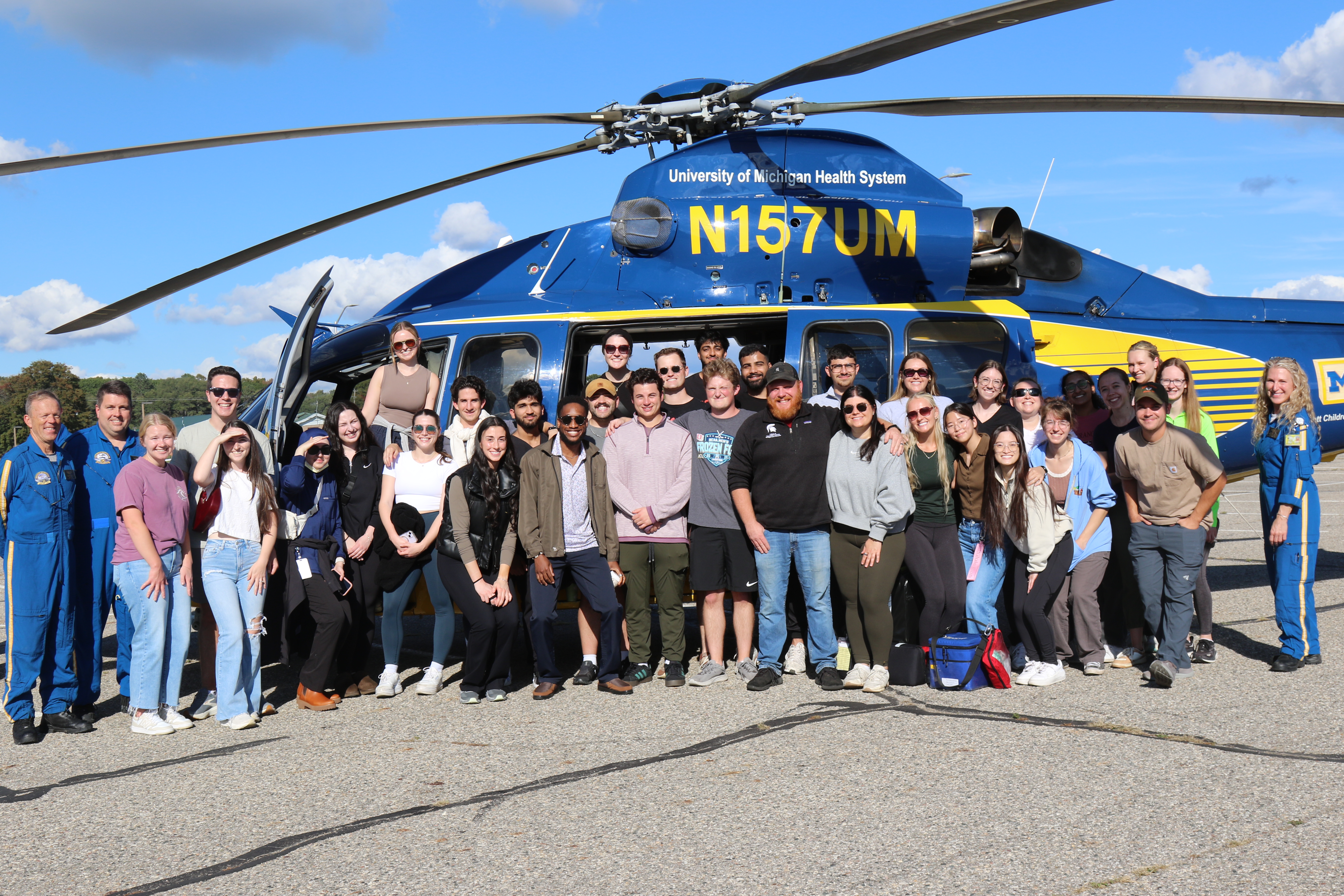 Group photo of students and instructors on the side of a landed blue and yellow helicopter from University of Michigan Health System.