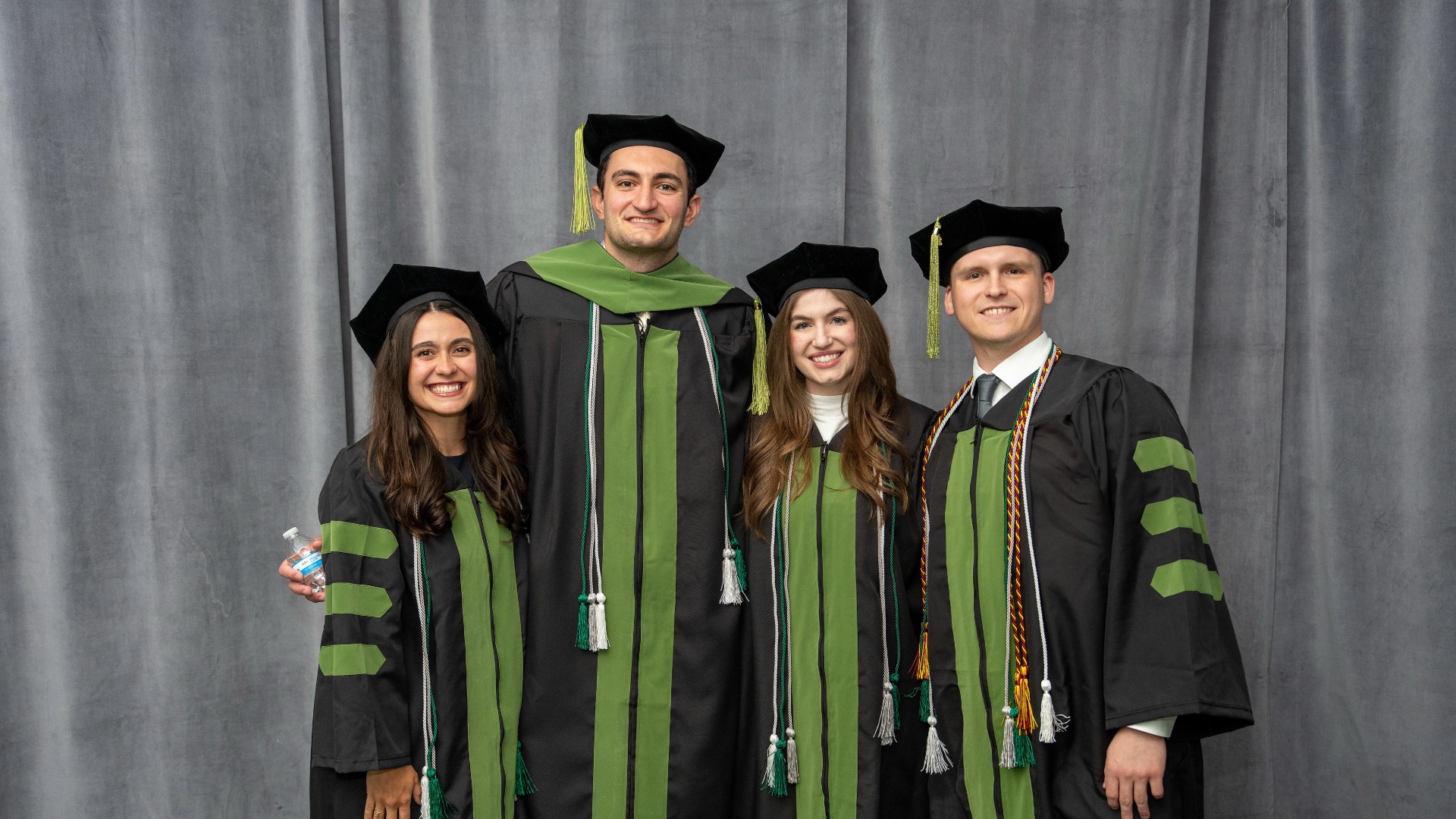 Four students in Michigan State University green doctoral regalia smiling in front of a grey backdrop