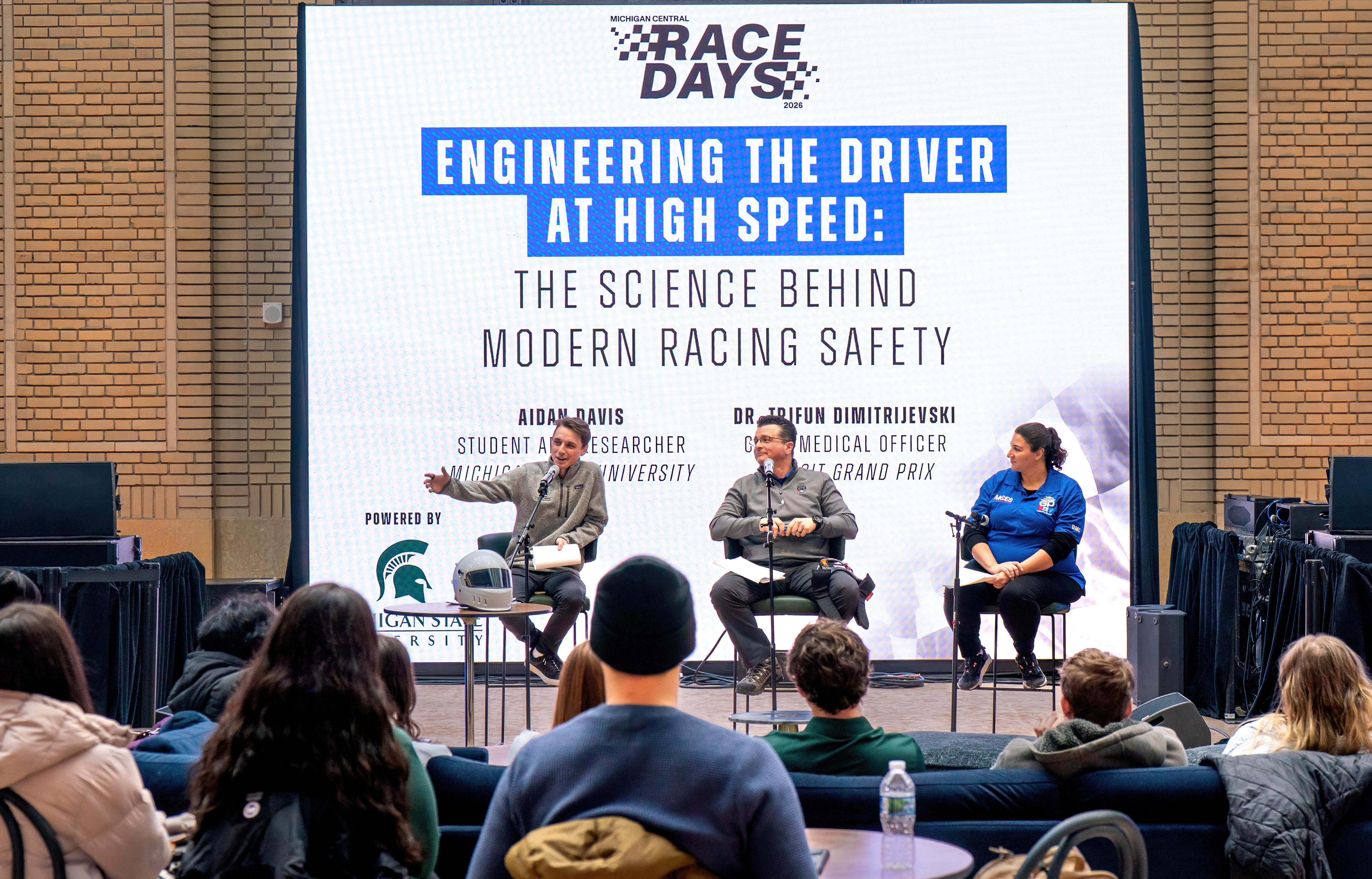 Three speakers on stage at an event discussing racing safety with an audience in attendance
