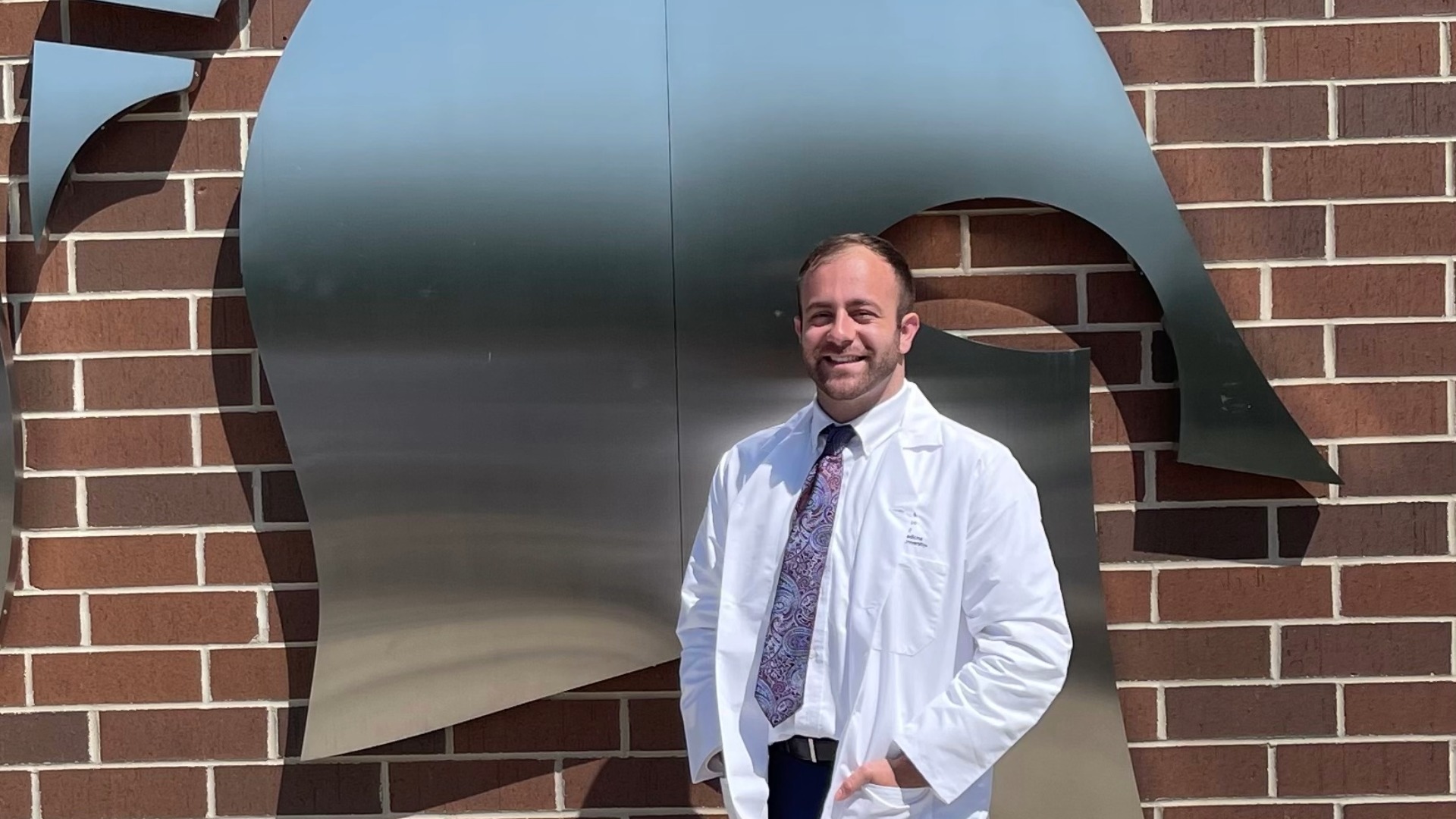 Alex Pasculle wearing a white doctors coat standing in front of a large stainless steel spartan helmet emblem at MSU Breslin Student Events Center