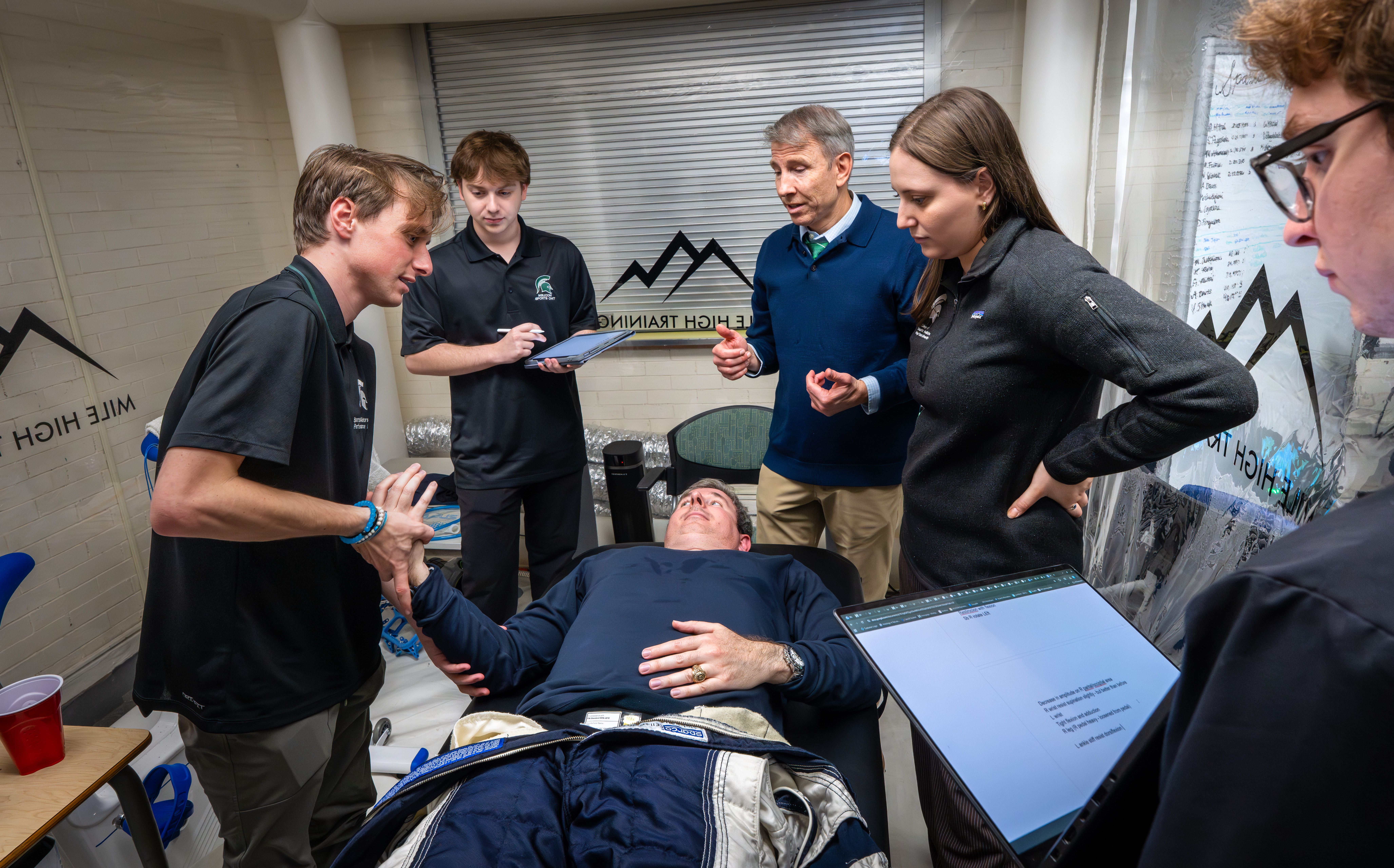 Spartan Performance Lab team of five surrounding a patient lying on an exam table with their right hand being evaluated using osteopathic manipulative medicine (OMM).