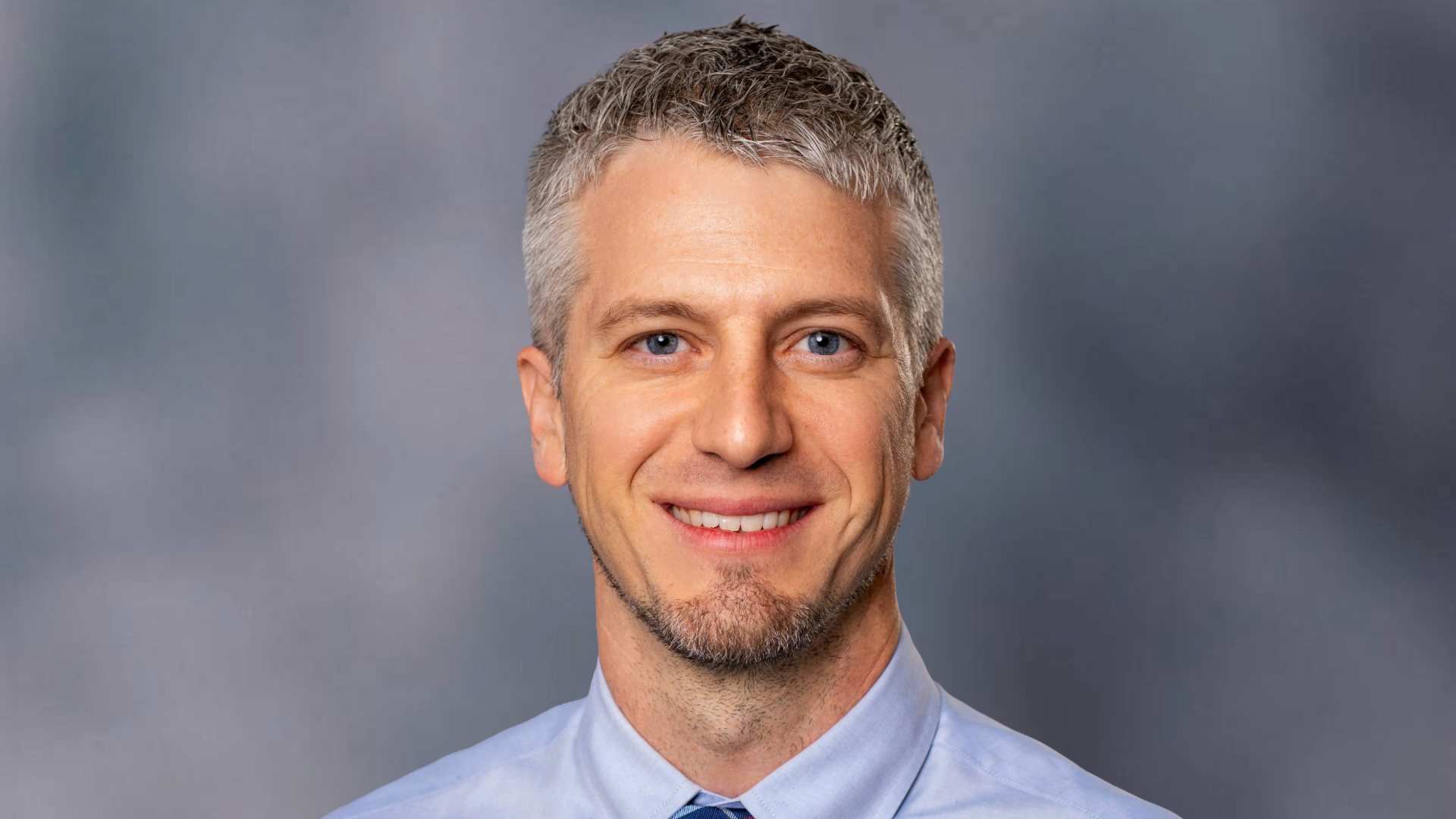 Headshot of Travis Gordon, D.O., smiling against a gray cloudy background, wearing a blue button-down shirt with a red and blue checkered tie.