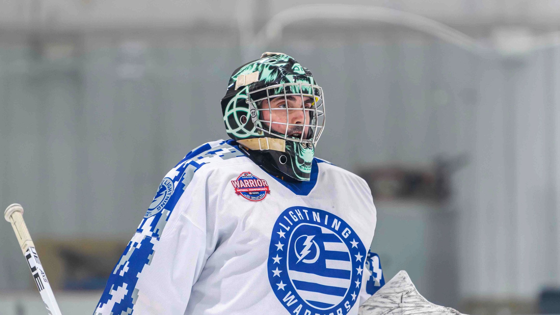 MSUCOM student Randall Nolin skating on the ice in a white hockey uniform, wearing a helmet and holding a hockey stick
