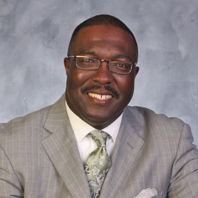 Headshot of Bob Kendrick smiling with square glasses in a checkered patterned grey suit and green floral tie with a white pocket square