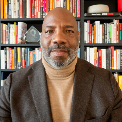 Headshot of Jelani Cobb soft smiling in a tan turtle neck sweater and black overcoat