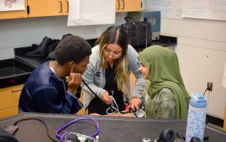 PA student helping two Cass Technical High School students take each others blood pressure. One student is wearing a stethoscope and the other is wearing a blood pressure cuff on their arm with the end of the stethoscope on their arm so the other student can hear their blood pressure.