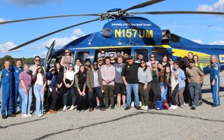 Group photo of students and instructors on the side of a landed blue and yellow helicopter from University of Michigan Health System.