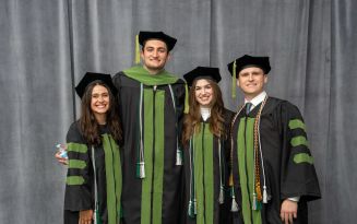 Four students in Michigan State University green doctoral regalia smiling in front of a grey backdrop