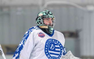 MSUCOM student Randall Nolin skating on the ice in a white hockey uniform, wearing a helmet and holding a hockey stick