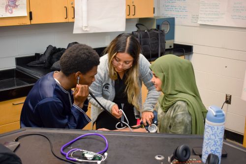 PA student helping two Cass Technical High School students take each others blood pressure. One student is wearing a stethoscope and the other is wearing a blood pressure cuff on their arm with the end of the stethoscope on their arm so the other student can hear their blood pressure.
